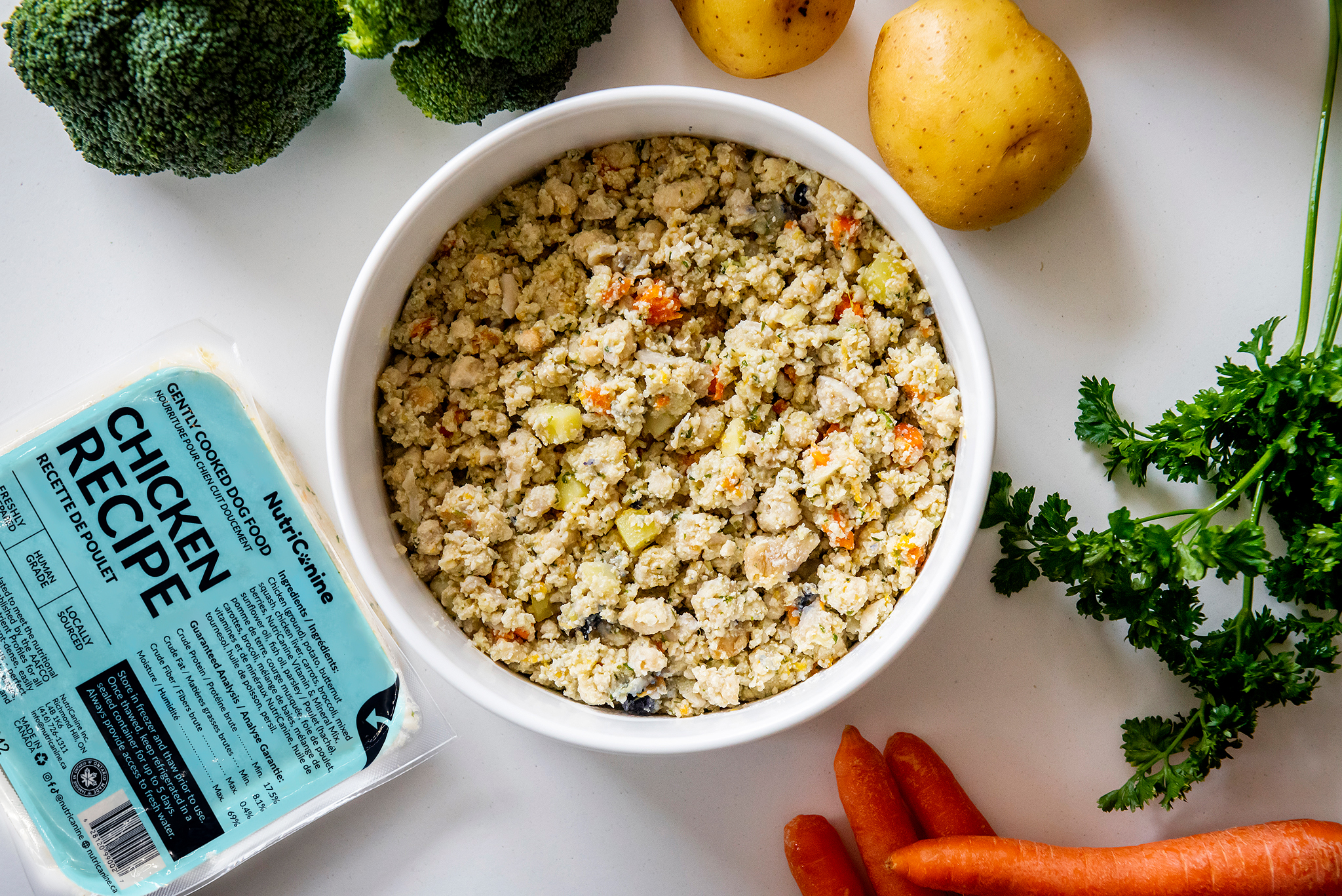 Bowl of cooked ground chicken with vegetables, surrounded by broccoli, potatoes, carrots, and parsley